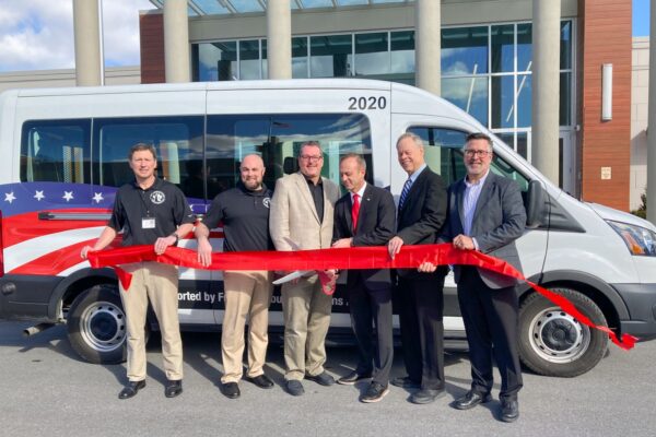 Six men stand in front of a van, holding a red ribbon. They appear to be at a ribbon-cutting event. The van features an American flag design.