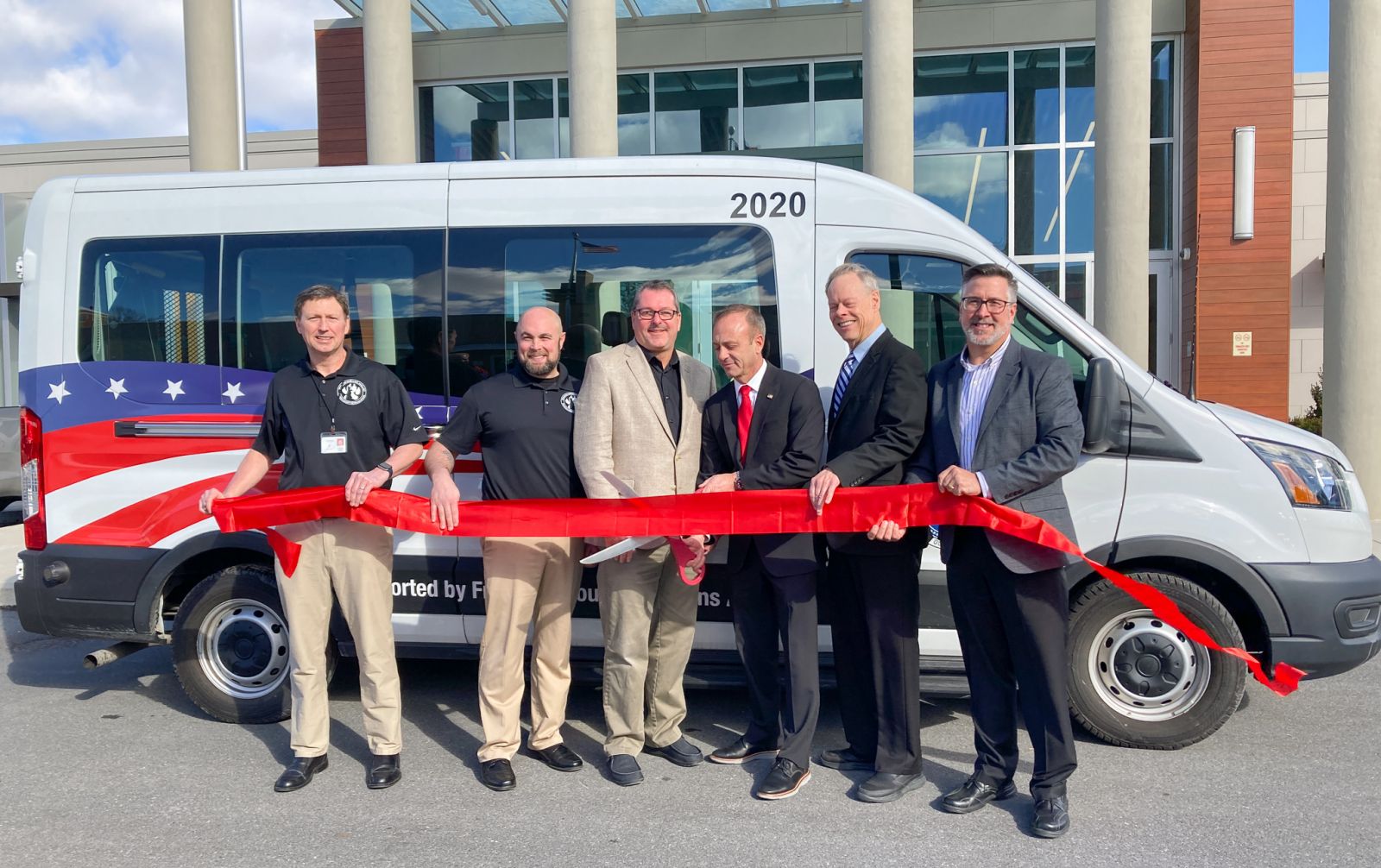 Six men stand in front of a van, holding a red ribbon. They appear to be at a ribbon-cutting event. The van features an American flag design.