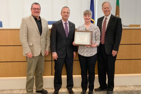 Four people standing, one woman holding a framed certificate. They are in a formal setting with flags and a wooden panel in the background.