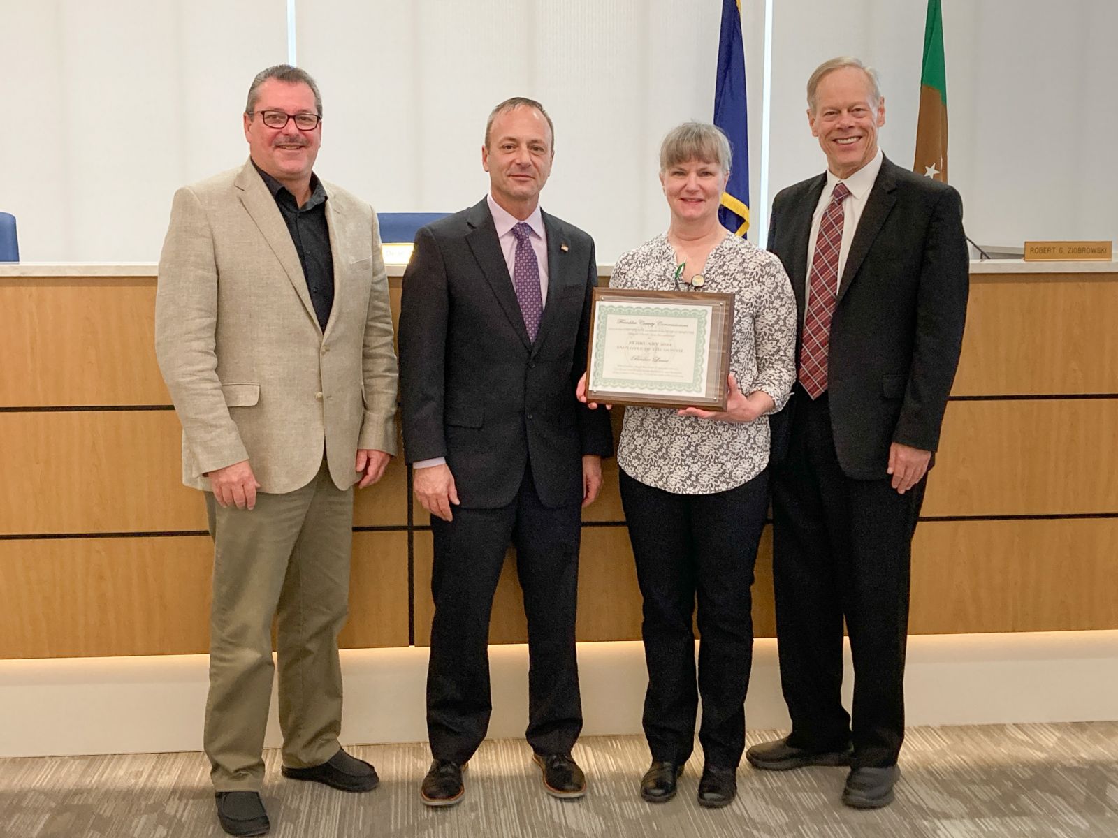 Four people standing, one woman holding a framed certificate. They are in a formal setting with flags and a wooden panel in the background.