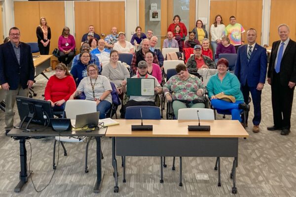 A group of people, including individuals in wheelchairs, pose together in a room with chairs and microphones at the front.