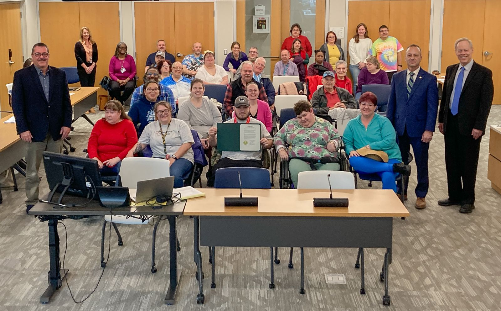 A group of people, including individuals in wheelchairs, pose together in a room with chairs and microphones at the front.