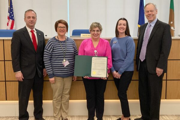 Five people standing together in an official setting, with one person holding a framed document. There are flags and a nameplate in the background.