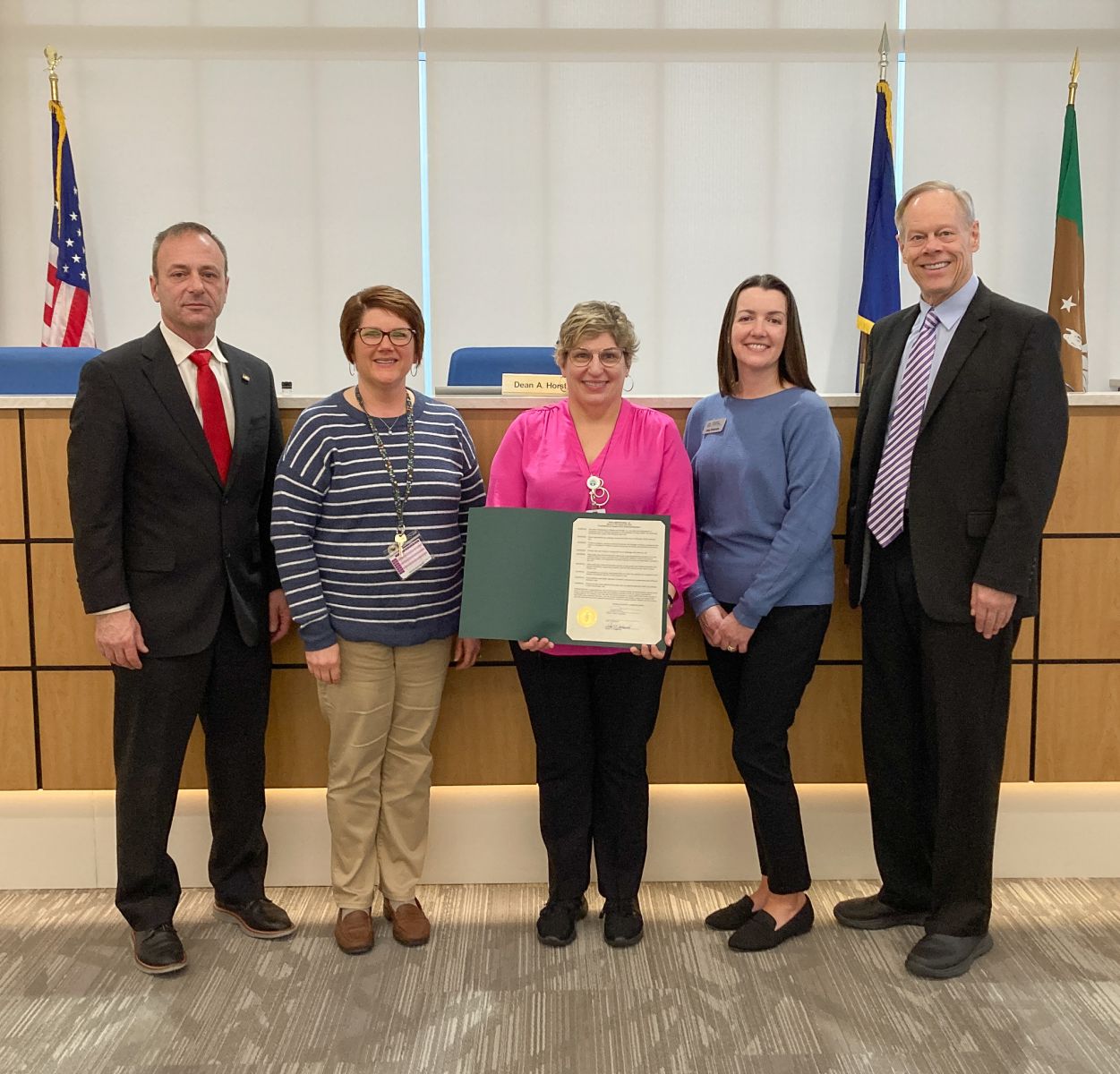 Five people standing together in an official setting, with one person holding a framed document. There are flags and a nameplate in the background.