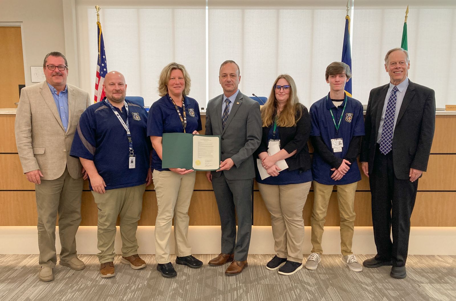 A group of seven people standing indoors, holding a certificate. Some are wearing matching blue uniforms. An American flag is visible in the background.