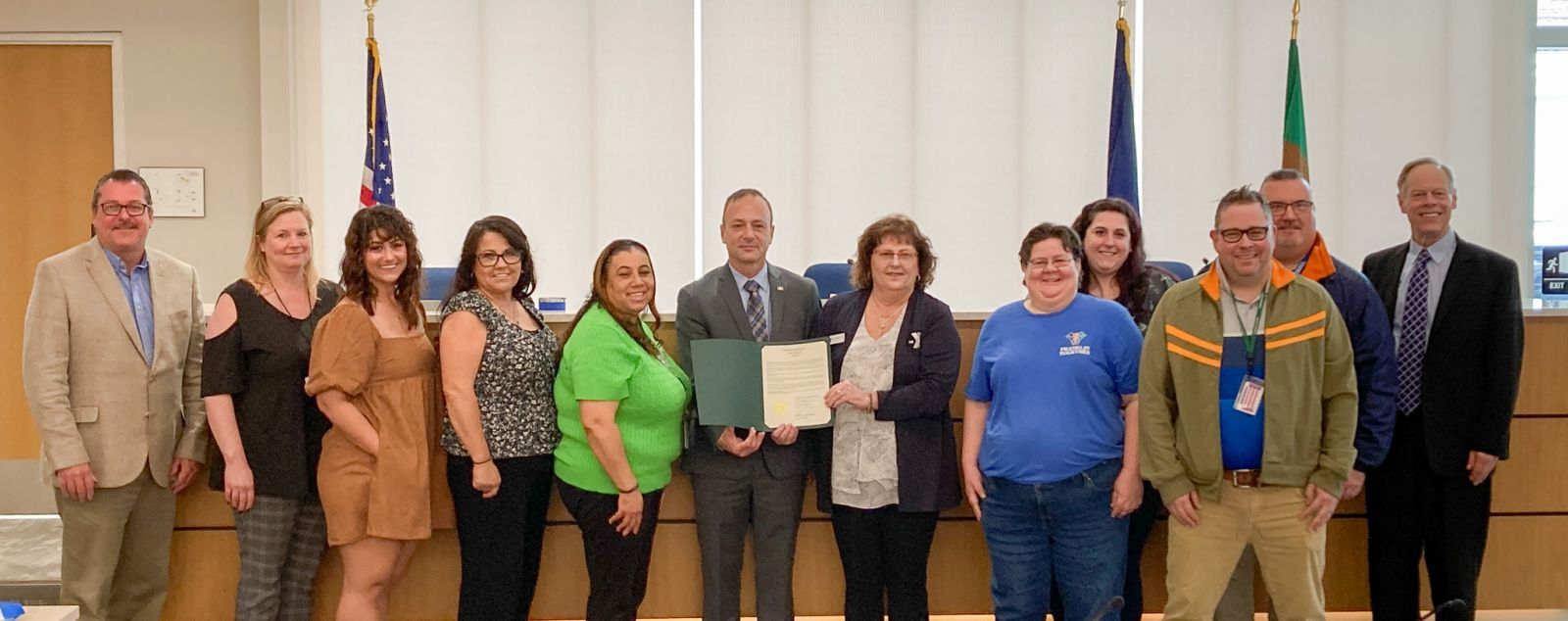 A group of people stands in front of a table. One person in the center holds a certificate. Flags are displayed in the background.