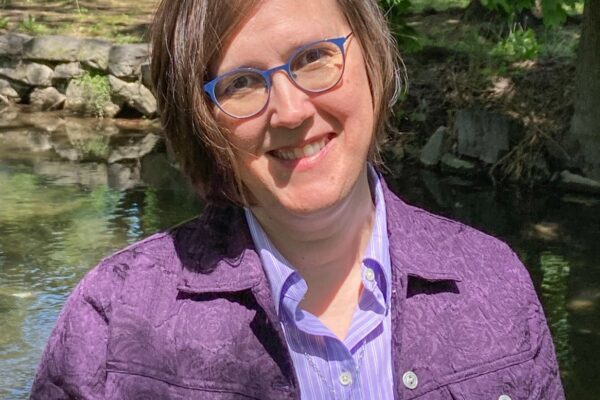 A person with short hair and glasses smiles while standing outdoors near a stone wall and trees, wearing a purple jacket and a striped shirt.