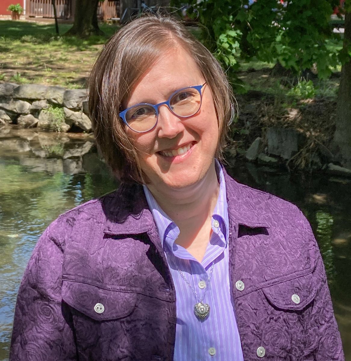 A person with short hair and glasses smiles while standing outdoors near a stone wall and trees, wearing a purple jacket and a striped shirt.