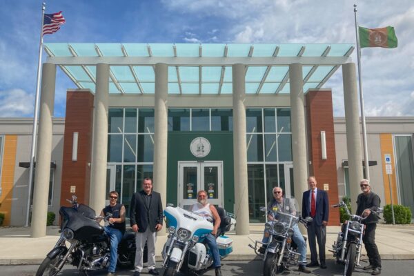 A group of five people standing in front of a building with three motorcycles parked nearby. Two flags are visible, one American and one green flag.