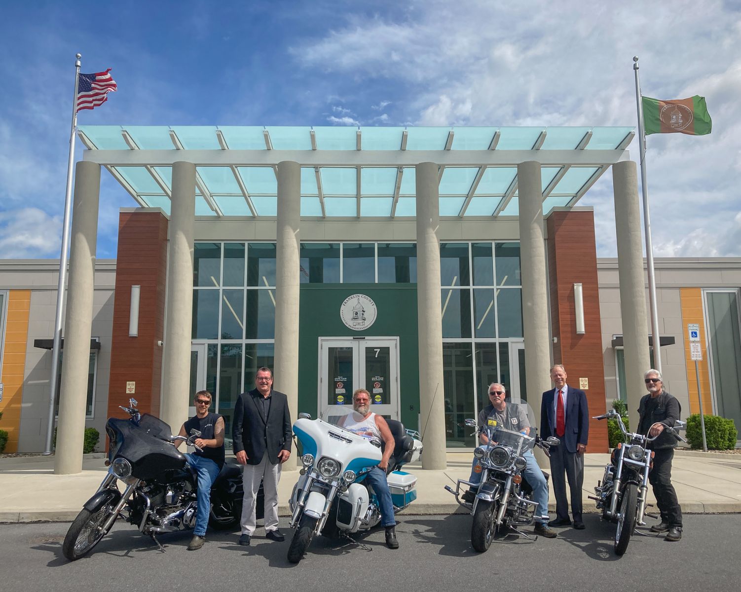 A group of five people standing in front of a building with three motorcycles parked nearby. Two flags are visible, one American and one green flag.