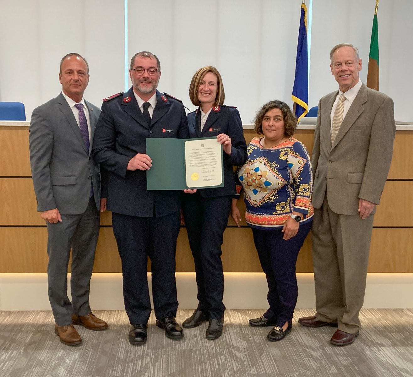 Five people standing in a row; two in uniforms hold a certificate. A flag and a podium are in the background.