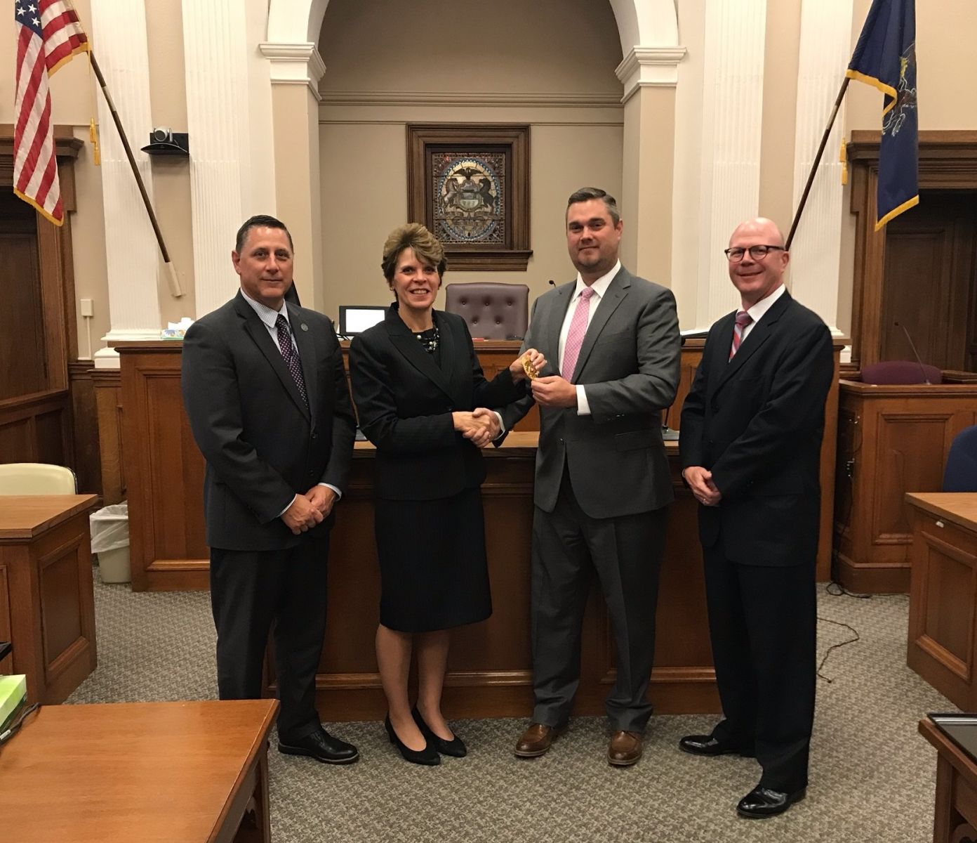 Four people in formal attire stand in a courtroom. One person is handing an object to another, who is smiling. Flags and a shield emblem are visible in the background.