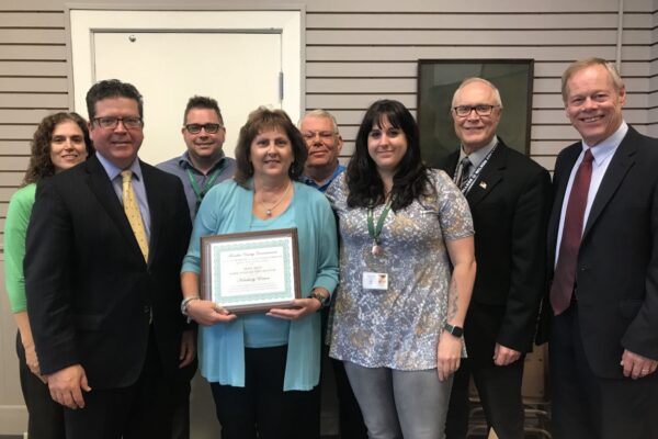 A group of eight people stands indoors, with one woman holding a framed certificate. Cropped blinds and framed certificate details in the background.