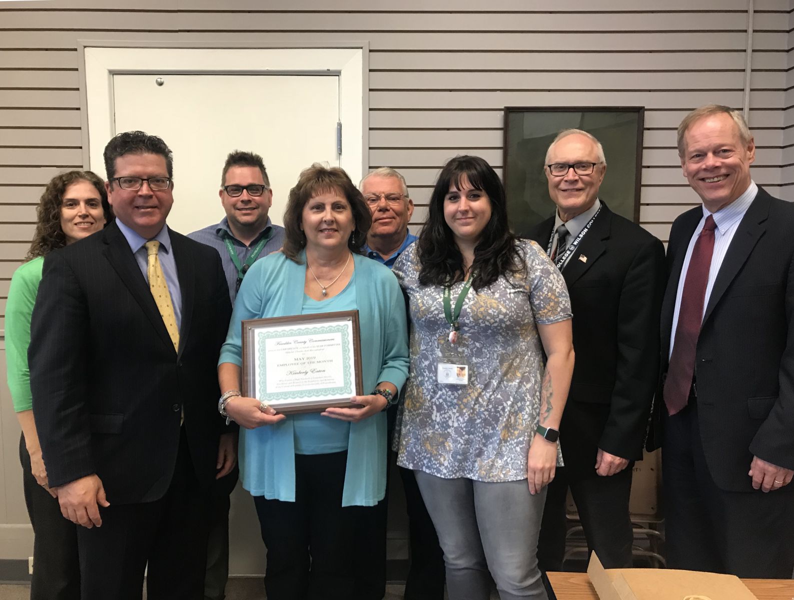 A group of eight people stands indoors, with one woman holding a framed certificate. Cropped blinds and framed certificate details in the background.