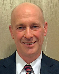 Bald man in a suit with a patterned tie, smiling against a textured, light-colored background.