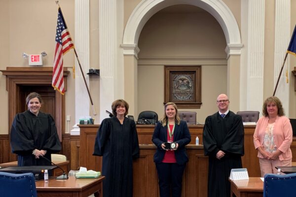 Five people in a courtroom, three in judicial robes and two in formal attire, standing in front of a judge’s bench with flags and wood-paneled walls in the background.