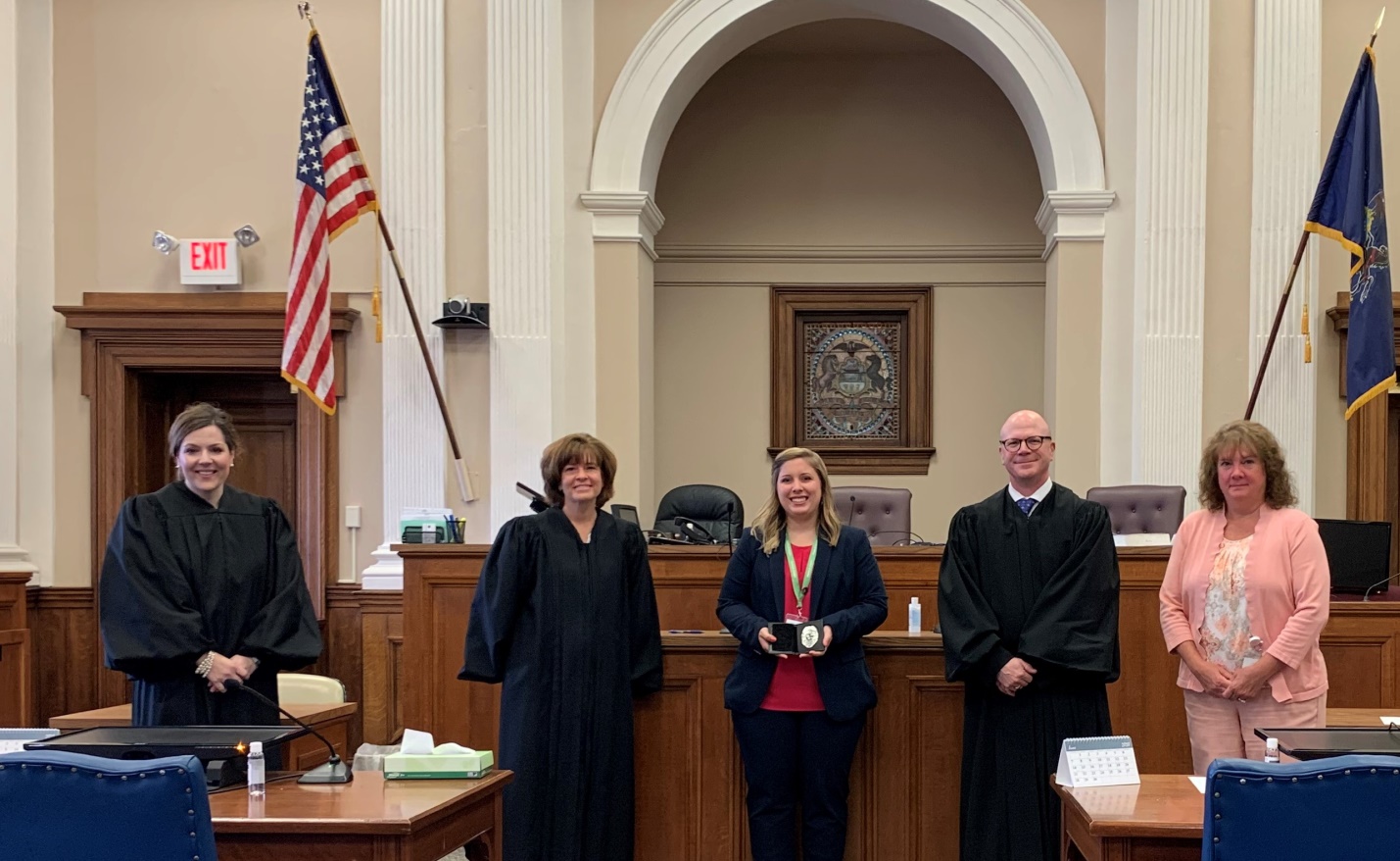 Five people in a courtroom, three in judicial robes and two in formal attire, standing in front of a judge’s bench with flags and wood-paneled walls in the background.