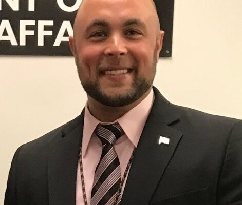 A man in a suit and tie smiles, standing indoors in front of a partial sign.