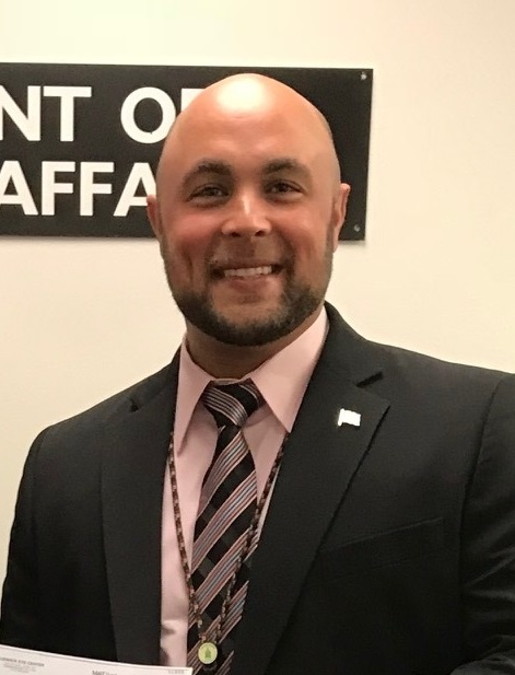 A man in a suit and tie smiles, standing indoors in front of a partial sign.