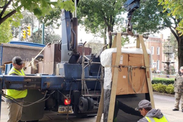 Workers in yellow vests use a crane to lift a large, packaged object on a city sidewalk. Trees and buildings are visible in the background.