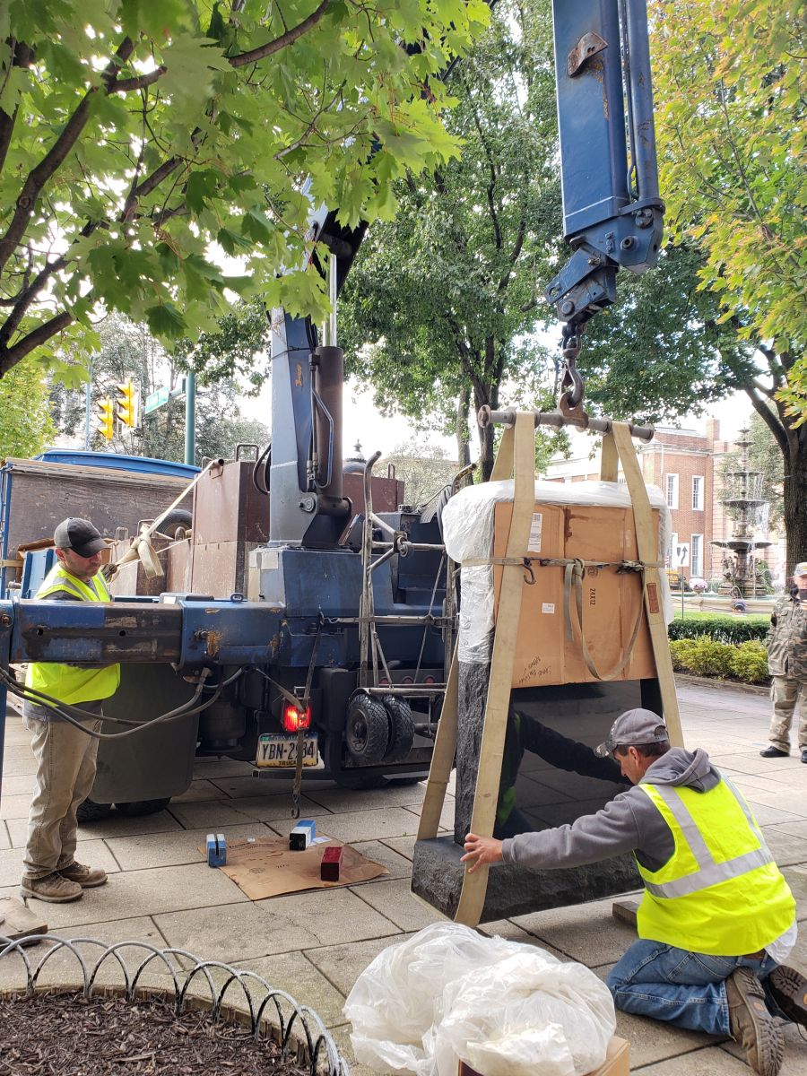 Workers in yellow vests use a crane to lift a large, packaged object on a city sidewalk. Trees and buildings are visible in the background.