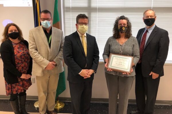 Five people stand in a room, four wearing masks. The woman in the center holds a framed certificate. They are in front of a window and flag.