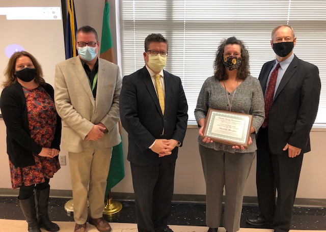 Five people stand in a room, four wearing masks. The woman in the center holds a framed certificate. They are in front of a window and flag.