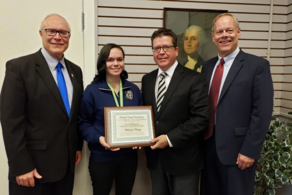 Four people stand indoors, one holding a framed certificate. They are all smiling. A portrait hangs on the wall in the background.