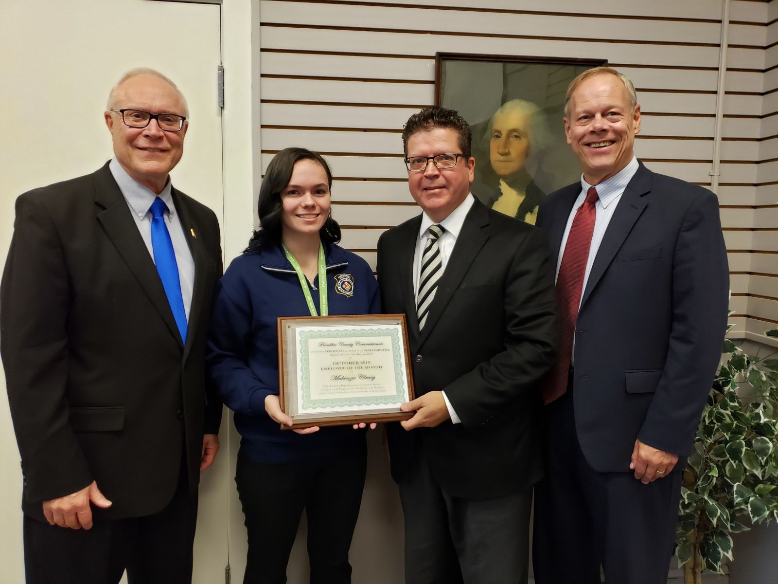 Four people stand indoors, one holding a framed certificate. They are all smiling. A portrait hangs on the wall in the background.