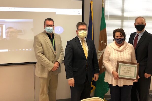 Four people in masks stand indoors; one holds a certificate. A webinar is displayed on a screen in the background. Flags are visible beside the participants.