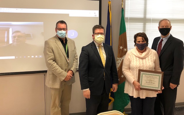 Four people in masks stand indoors; one holds a certificate. A webinar is displayed on a screen in the background. Flags are visible beside the participants.