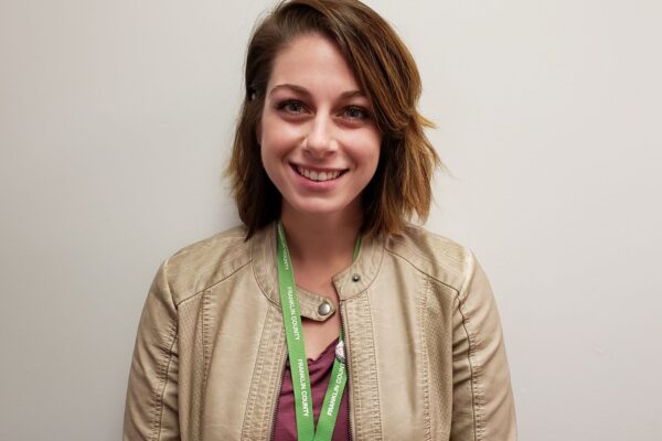 A person with shoulder-length brown hair wearing a beige jacket and a lanyard, standing against a plain background, smiling at the camera.