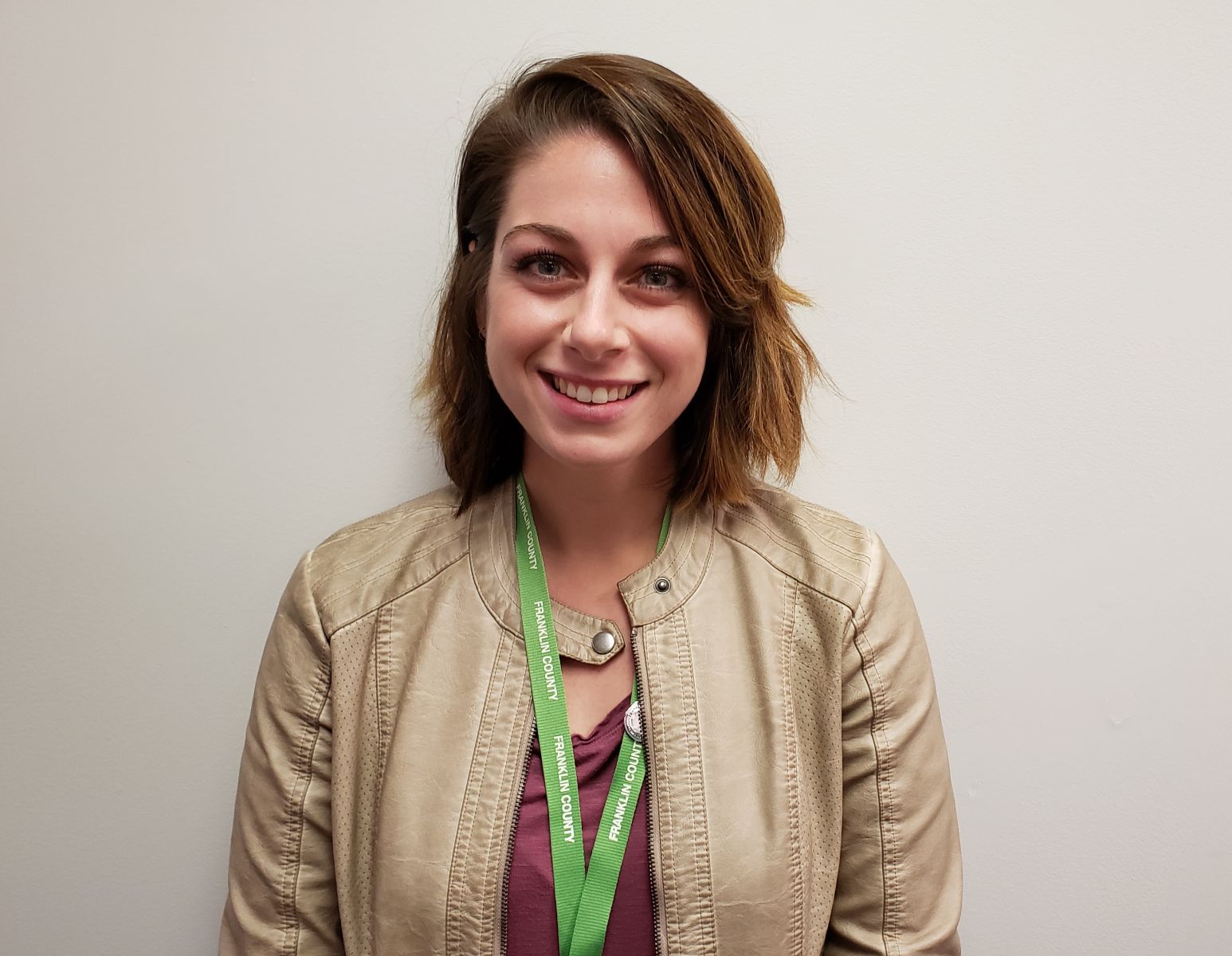 A person with shoulder-length brown hair wearing a beige jacket and a lanyard, standing against a plain background, smiling at the camera.