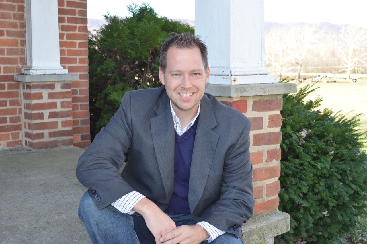 A person in a suit jacket and jeans smiles, crouching in front of a brick column. Bushes and a grassy area are in the background.