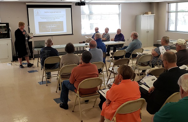 A group of people in a classroom setting listens to a presentation about new equipment by a speaker in front of a projector screen.