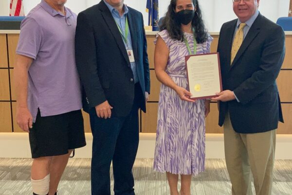 Four people standing indoors. One person holds a framed certificate. A man on the left wears a purple shirt and has a medical boot. U.S. flags and a wood podium are in the background.