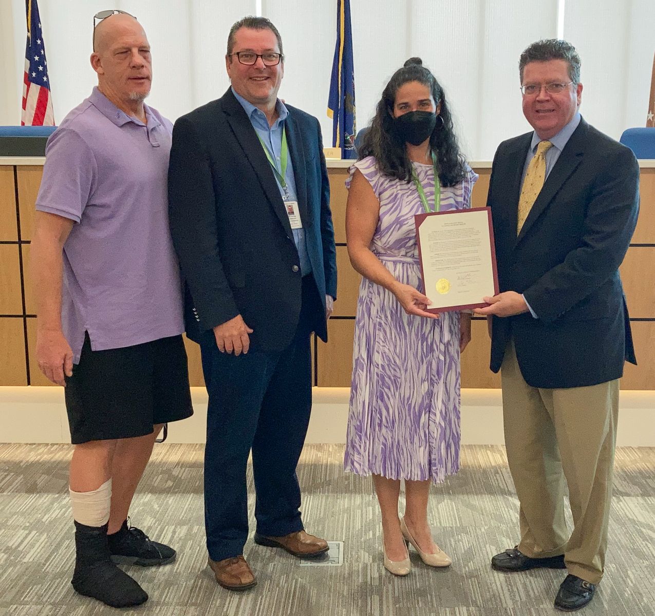 Four people standing indoors. One person holds a framed certificate. A man on the left wears a purple shirt and has a medical boot. U.S. flags and a wood podium are in the background.