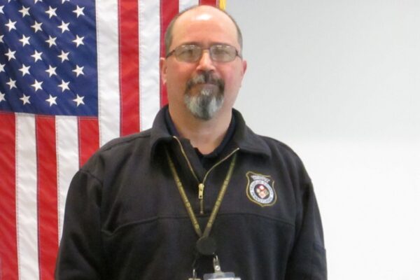 A man with glasses and a goatee stands in front of an American flag, wearing a black jacket with an insignia and ID badge.