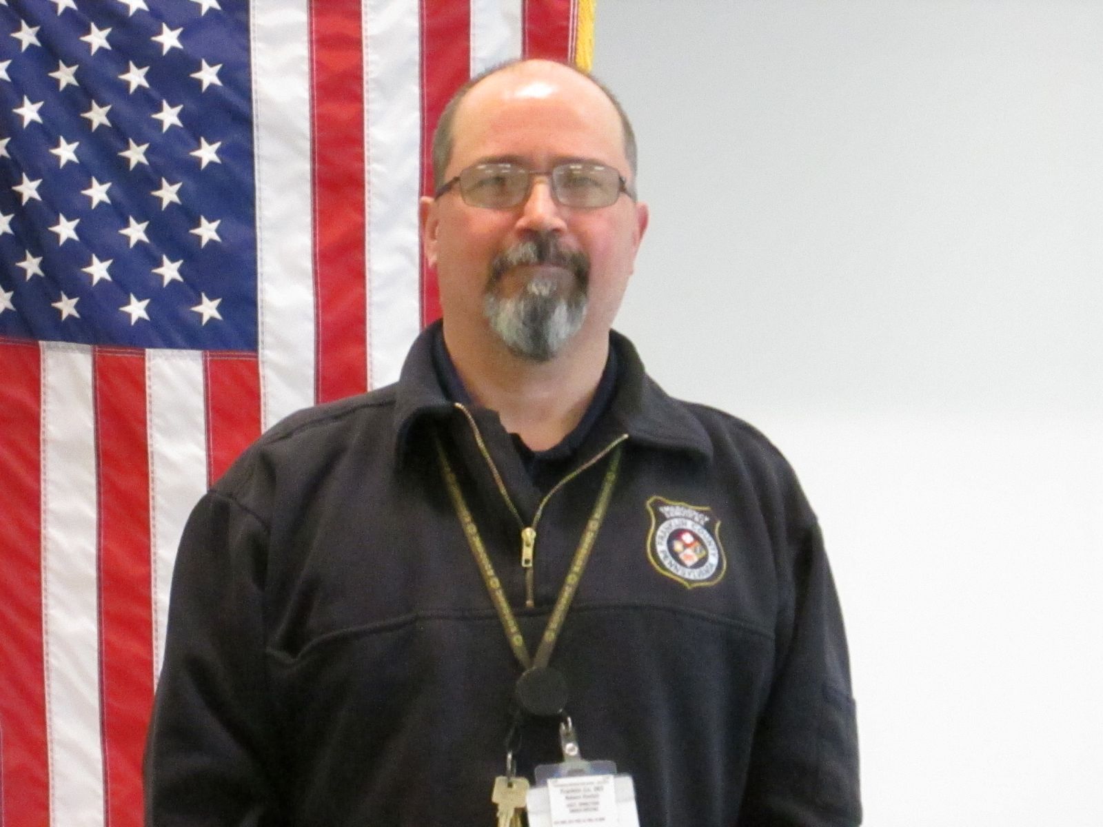 A man with glasses and a goatee stands in front of an American flag, wearing a black jacket with an insignia and ID badge.