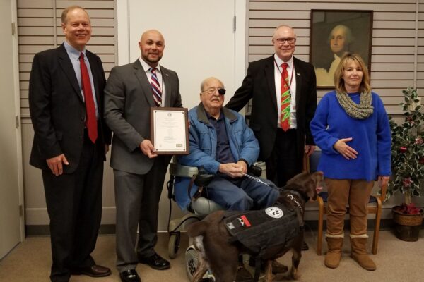 A group of five people, including a person in a wheelchair, pose with a certificate and a service dog in an indoor setting.