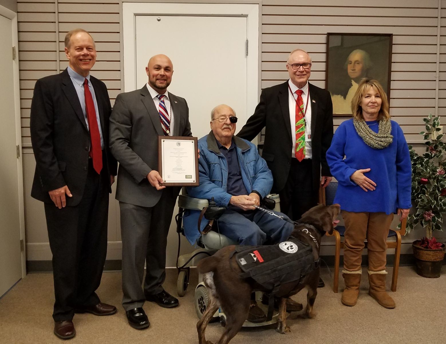 A group of five people, including a person in a wheelchair, pose with a certificate and a service dog in an indoor setting.