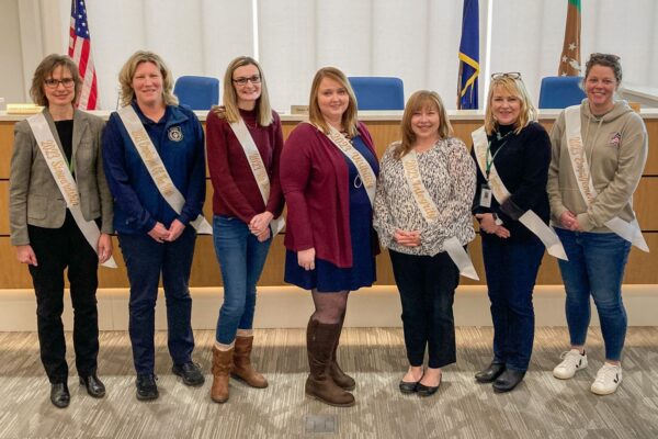 Seven women wearing sashes stand in a row in a room with flags.