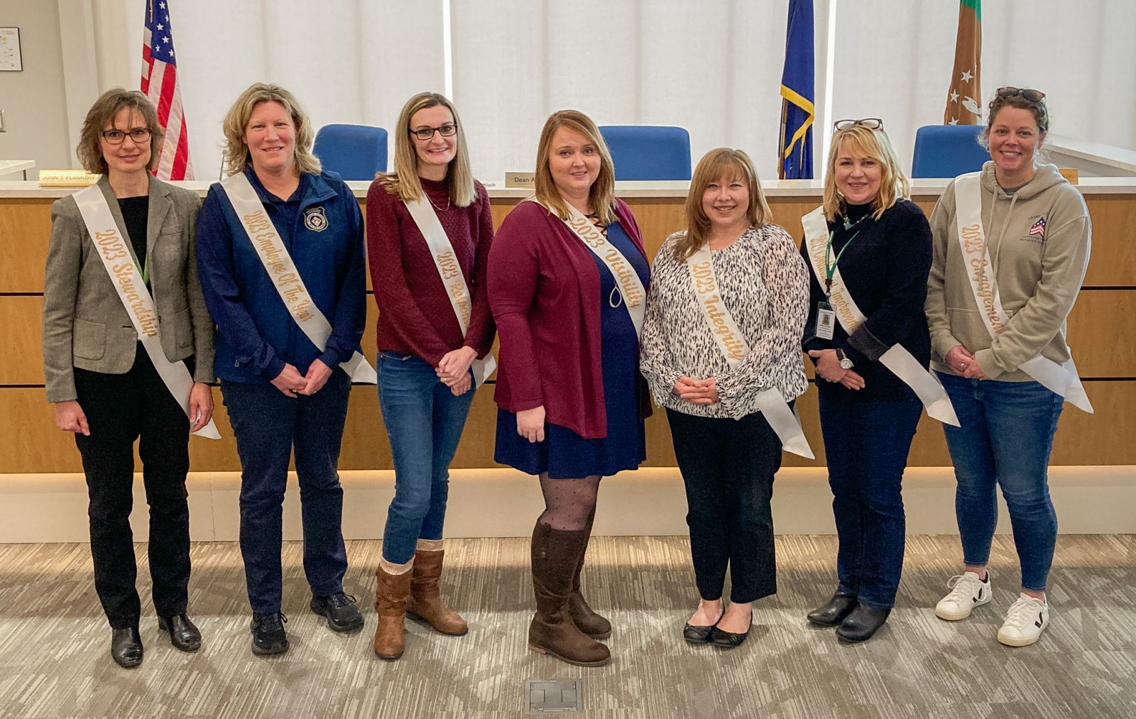 Seven women wearing sashes stand in a row in a room with flags.