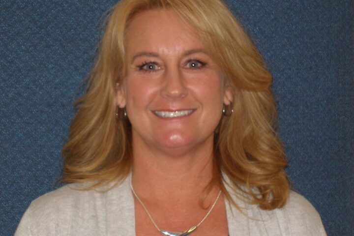 A person with shoulder-length blonde hair smiles against a textured blue background, wearing a light gray cardigan, dark top, and a silver necklace.