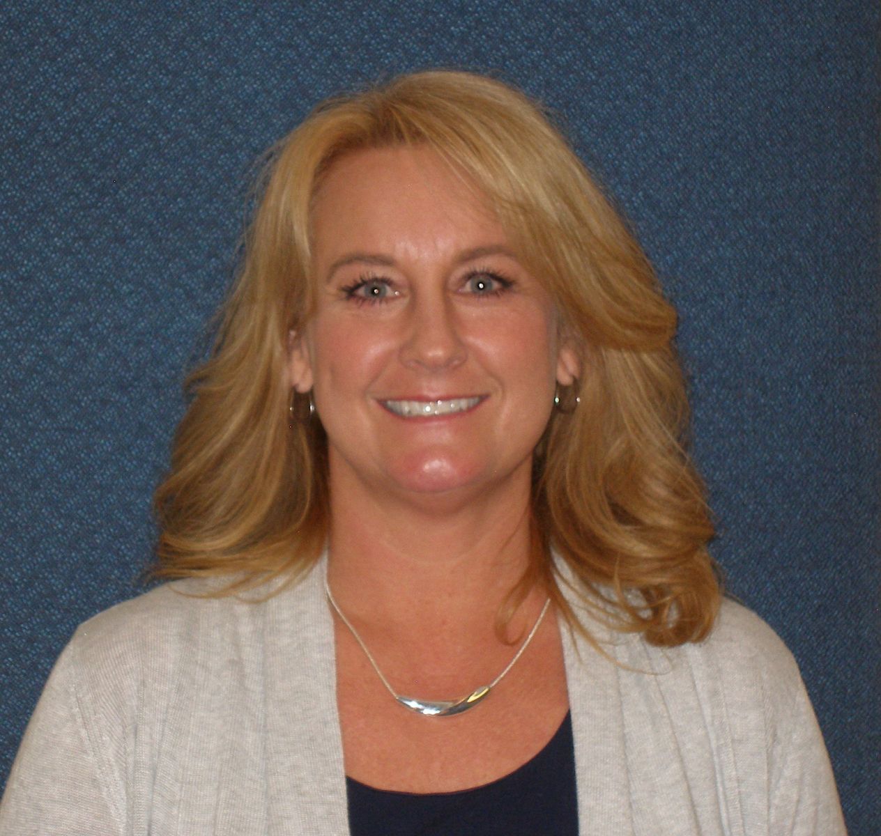 A person with shoulder-length blonde hair smiles against a textured blue background, wearing a light gray cardigan, dark top, and a silver necklace.