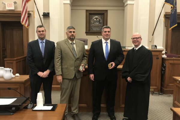 Four men in formal attire, including a judge in robes, stand in a courtroom. Two men hold plaques. An American flag and courtroom decor are visible in the background.