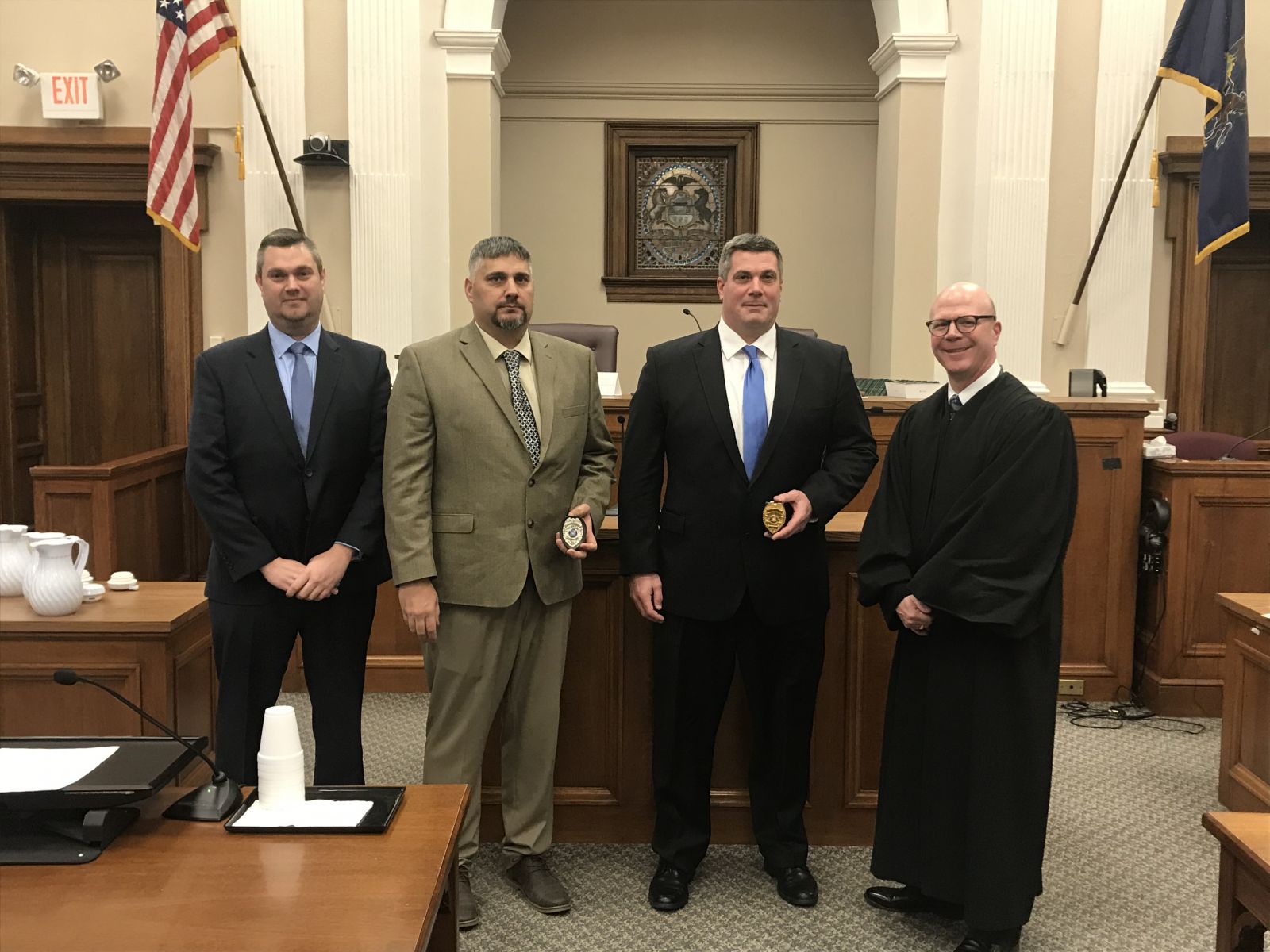 Four men in formal attire, including a judge in robes, stand in a courtroom. Two men hold plaques. An American flag and courtroom decor are visible in the background.