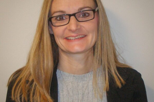 A person with long blonde hair, wearing glasses, a black blazer, and a gray top stands against a plain background.