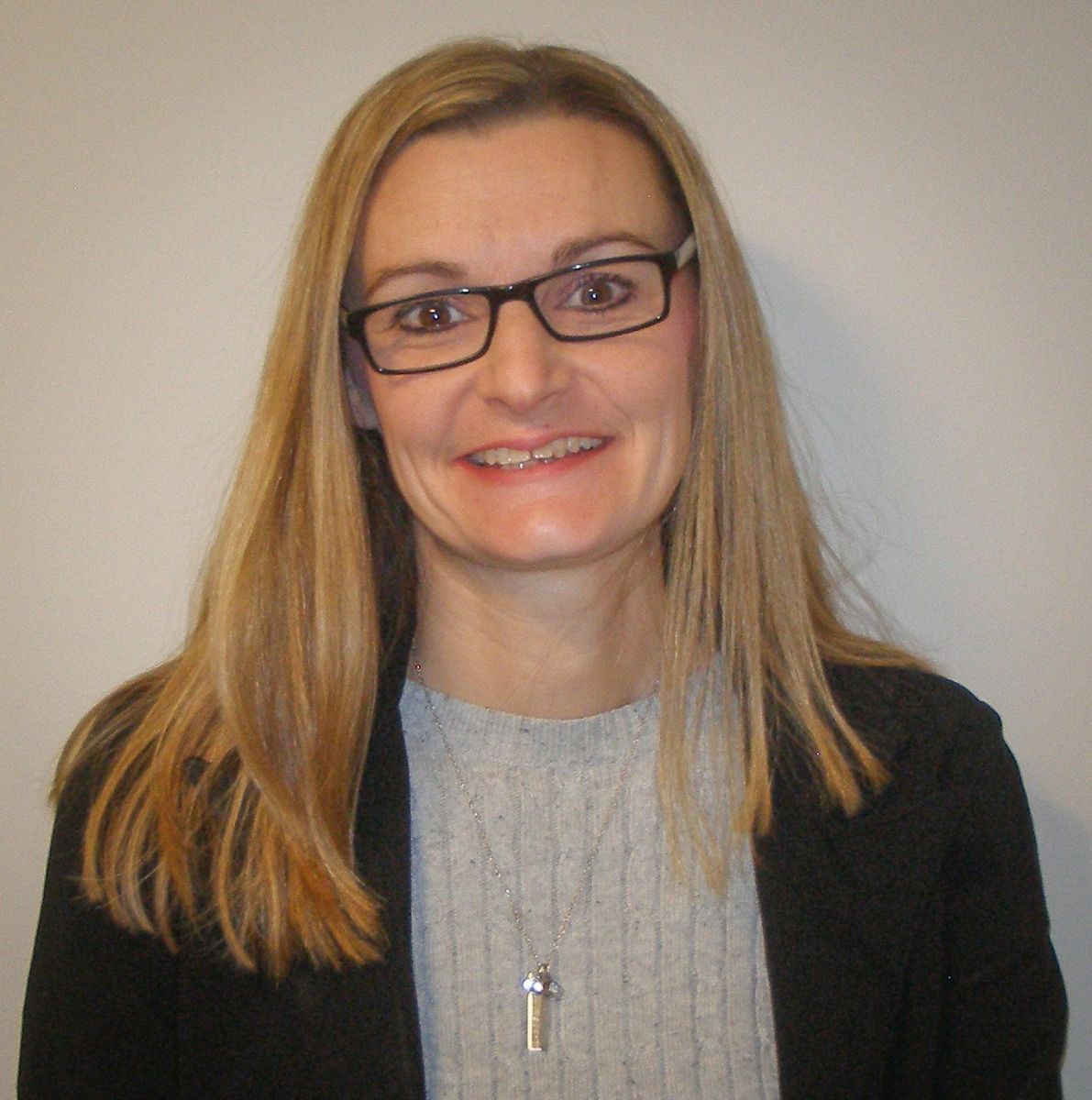 A person with long blonde hair, wearing glasses, a black blazer, and a gray top stands against a plain background.