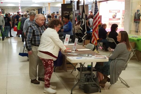 People interact at tables in a busy indoor event space, with informational displays and an American flag visible.